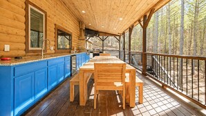 A wooden cabin with a covered porch, a dining table, and blue cabinets. - Sleeps 28! • Loaded w/ Amenities • Mins from Lake! (Broken Bow)