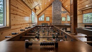 A spacious indoor area with wooden walls and a stone fireplace, featuring a foosball table and weightlifting equipment. - Sleeps 28! • Loaded w/ Amenities • Mins from Lake! (Broken Bow)