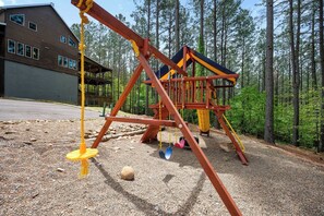 A playground with a swing set and a climbing structure, surrounded by trees and a building in the background. - Sleeps 28! • Loaded w/ Amenities • Mins from Lake! (Broken Bow)