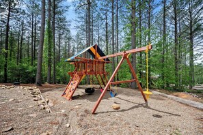 A playground with a wooden structure, including a slide and swings, set in a forested area with gravel ground. - Sleeps 28! • Loaded w/ Amenities • Mins from Lake! (Broken Bow)