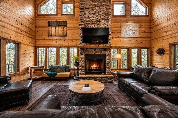 A log cabin interior with a stone fireplace, leather furniture, and large windows.