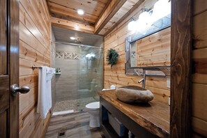 A wooden cabin bathroom with a glass-enclosed shower, a white toilet, and a stone sink. - Sleeps 28! • Loaded w/ Amenities • Mins from Lake! (Broken Bow)
