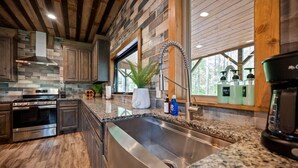 A modern kitchen with granite countertops, stainless steel appliances, and a large window offering a view of the outdoors. - Sleeps 28! • Loaded w/ Amenities • Mins from Lake! (Broken Bow)