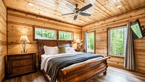 A wooden cabin bedroom with a bed, nightstands, a ceiling fan, and large windows. - Sleeps 28! • Loaded w/ Amenities • Mins from Lake! (Broken Bow)
