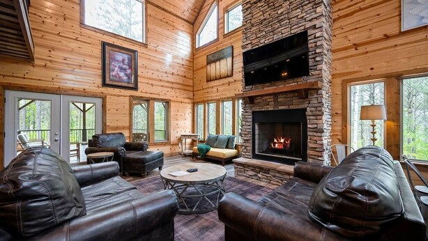 A log cabin interior with a stone fireplace, leather sofas, and a large window overlooking the forest. - Sleeps 28! • Loaded w/ Amenities • Mins from Lake! (Broken Bow)