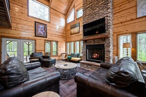 Een blokhut interieur met een stenen open haard, leren banken en een groot raam met uitzicht op het bos.