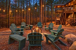 A forest setting with a fire pit surrounded by Adirondack chairs, a wooden deck, and a cabin structure.