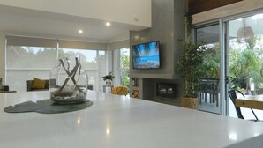Un salon moderne avec une grande table blanche, une cheminée, un téléviseur et une vue sur l’extérieur.