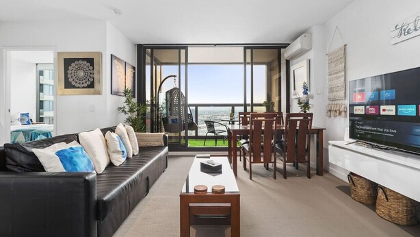 A modern living room with a sofa, coffee table, dining area, and a TV displaying streaming services. - Water Views Melbourne CBD - WiFi - Netflix (Melbourne)