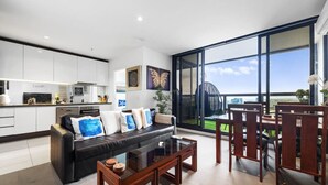 A modern living room with a sofa, coffee table, and dining area with chairs. There is a large window with a view of the city. - Water Views Melbourne CBD - WiFi - Netflix (Melbourne)