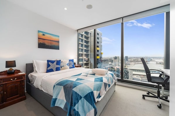 A bedroom with a large bed, a nightstand, a desk, and a chair. There is a large window with a view of buildings and a bridge.