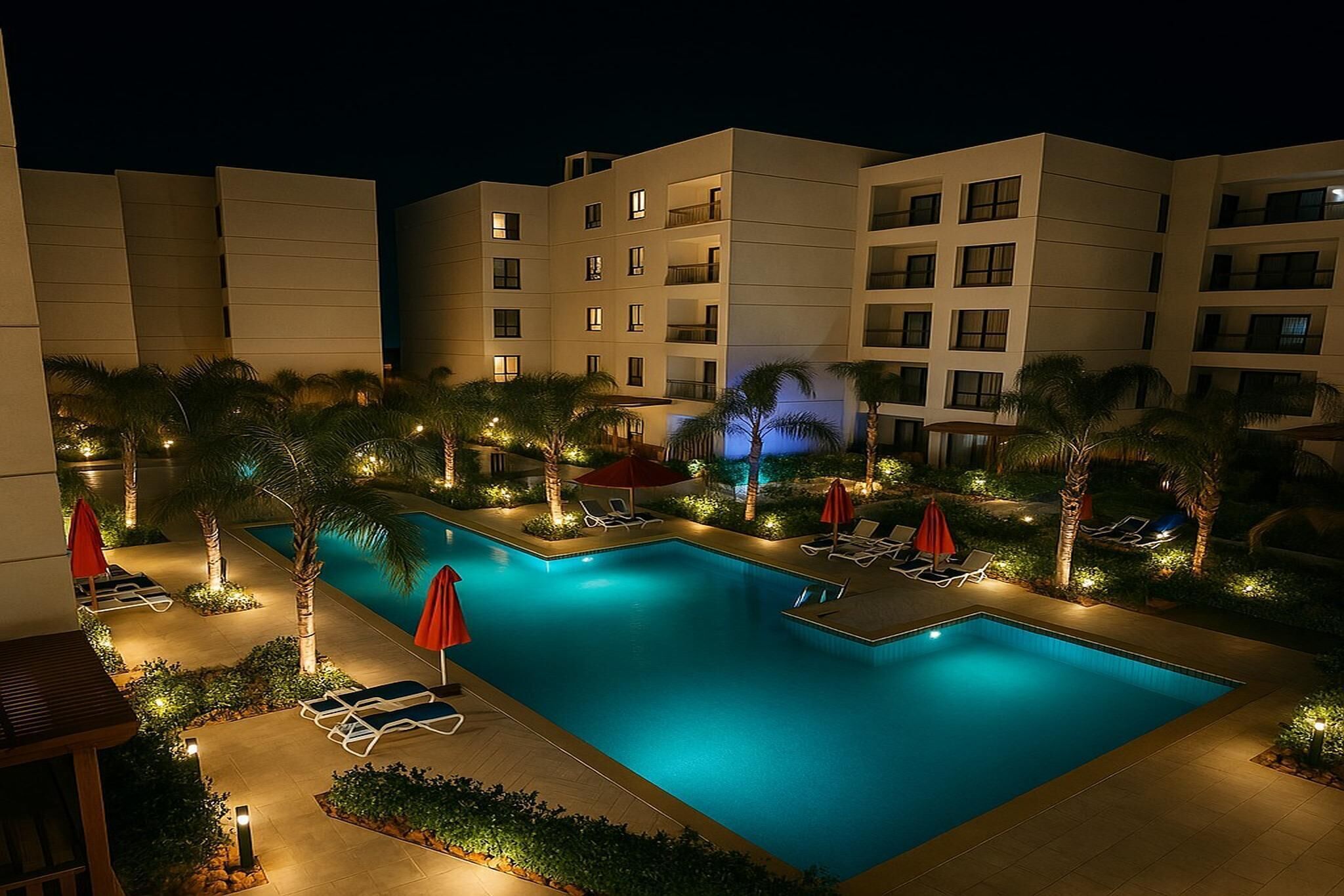 A nighttime view of a hotel pool area with a rectangular pool, lounge chairs, and palm trees.