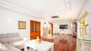 A spacious living room with a sofa, a white coffee table, a fireplace, and a TV mounted on the wall.
