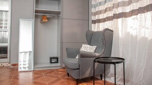 A room with a grey armchair, a small table, a shelving unit, and a window with curtains. - Full AirCon- 3Bed Villa- A/c- WiFi- HotWater (Freetown)