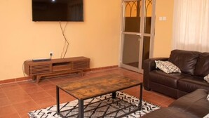A living room with a flat-screen TV mounted on the wall, a wooden entertainment unit, a brown leather sofa, and a patterned rug. - Full AirCon- 3Bed Villa- A/c- WiFi- HotWater (Freetown)