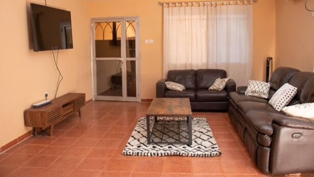 A living room with a sofa, a coffee table, a TV mounted on the wall, and a sliding glass door leading to a balcony. - Full AirCon- 3Bed Villa- A/c- WiFi- HotWater (Freetown)
