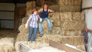 Two people sitting on hay bales inside a barn with corrugated metal walls and a metal roof.