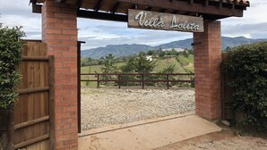 Una entrada con pérgola de madera, un letrero que dice "Villa Lolitá" y vista de un paisaje rural con montañas al fondo.