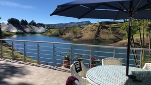 Una terraza con vistas a un lago y colinas, con una mesa y sillas bajo un paraguas grande.