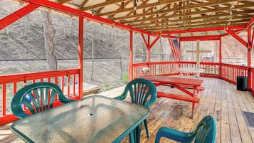 A covered outdoor deck with a table and chairs, wooden railings, and a view of a wooded area.