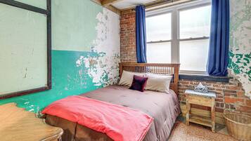 A room with a bed, a wooden headboard, a small table, a wicker basket, and a window with blue curtains.