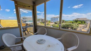 Área externa coberta com uma mesa redonda e duas cadeiras, janelas amplas com vista para casas e vegetação.