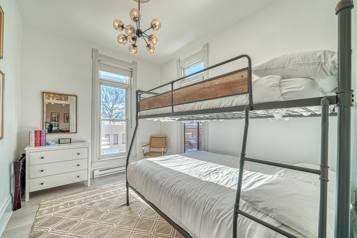 Une chambre avec un lit superposé doté d’une tête de lit en bois, une commode blanche, un miroir et un tapis à motifs.