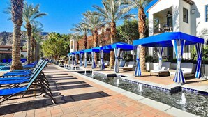Un espace aménagé au bord de la piscine, avec des chaises longues bleues et des parasols, des palmiers, et un bâtiment à la façade blanche.