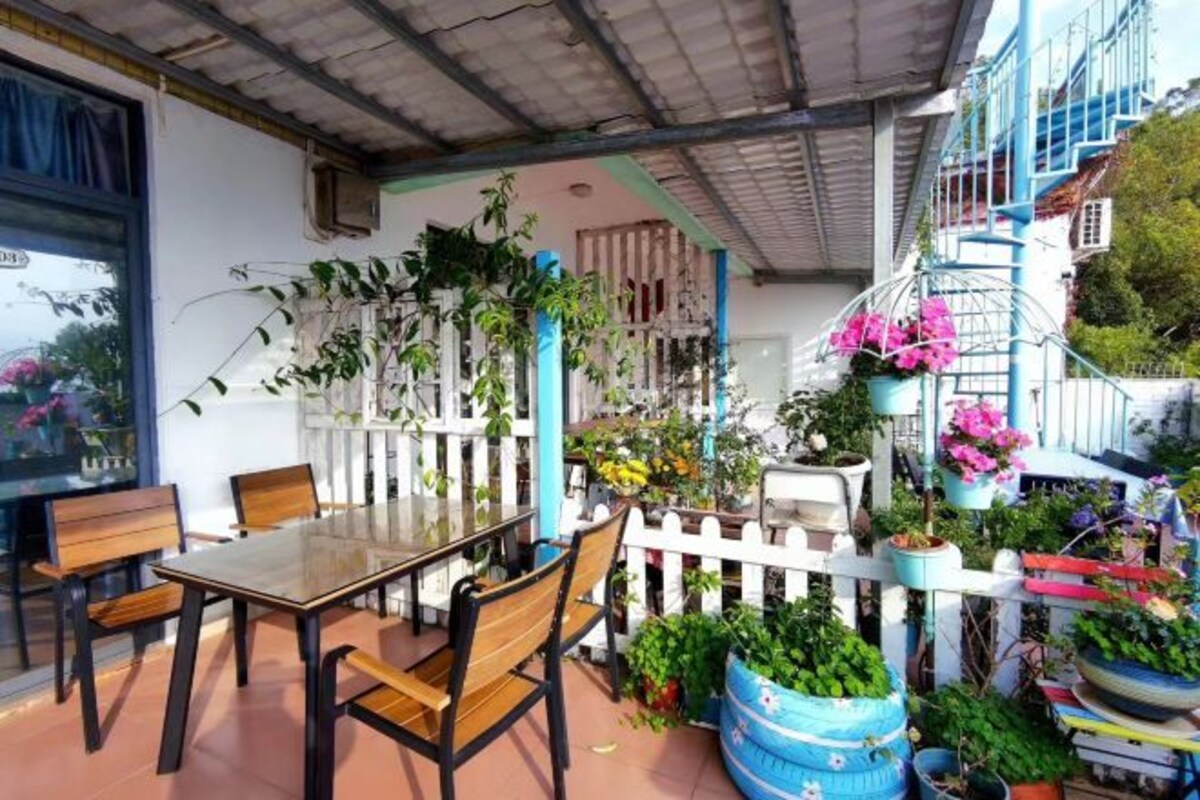 Un patio cubierto con una mesa de vidrio y sillas de madera, rodeado de plantas en macetas y una cerca blanca de piquetes.