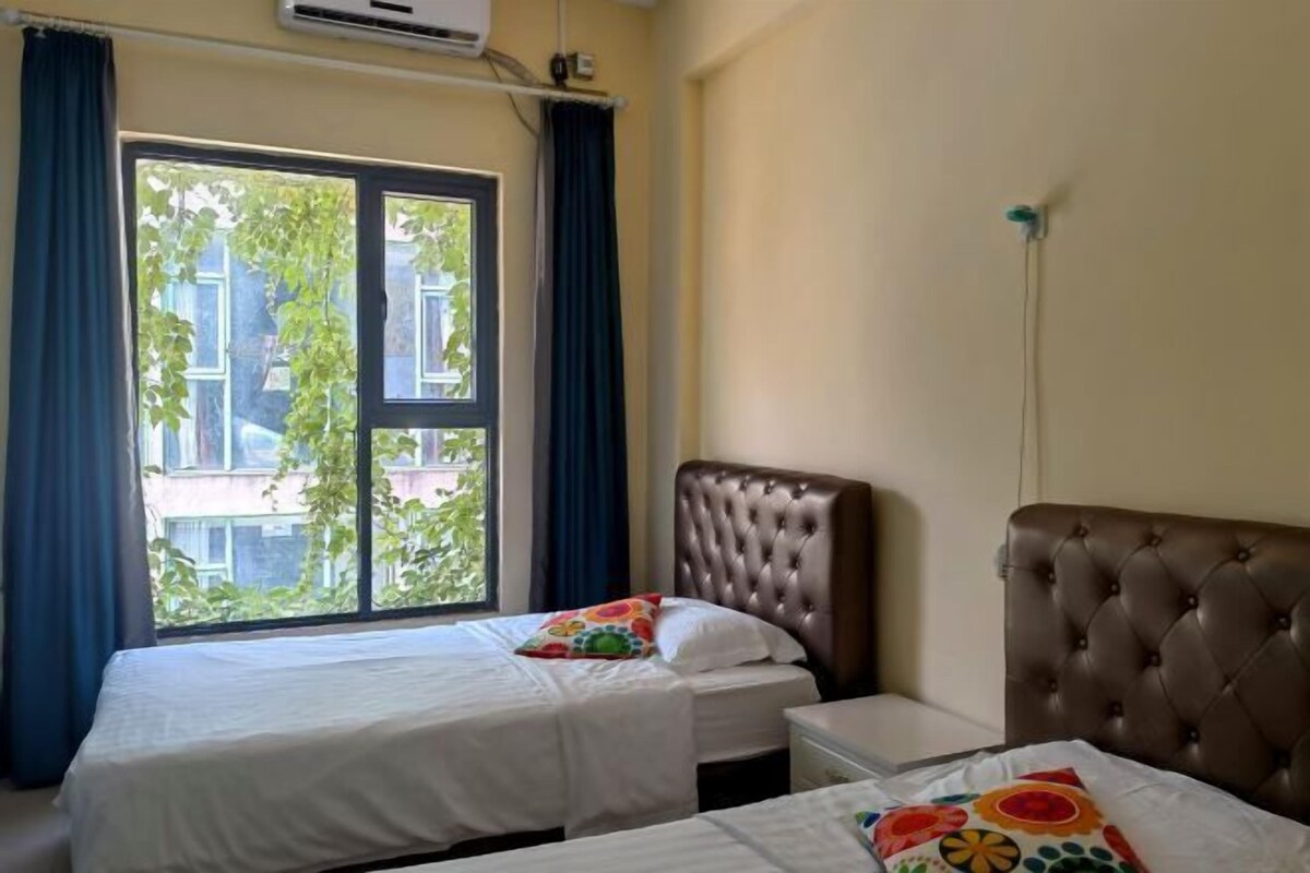A hotel room with two beds, a window with curtains, and an air conditioning unit.