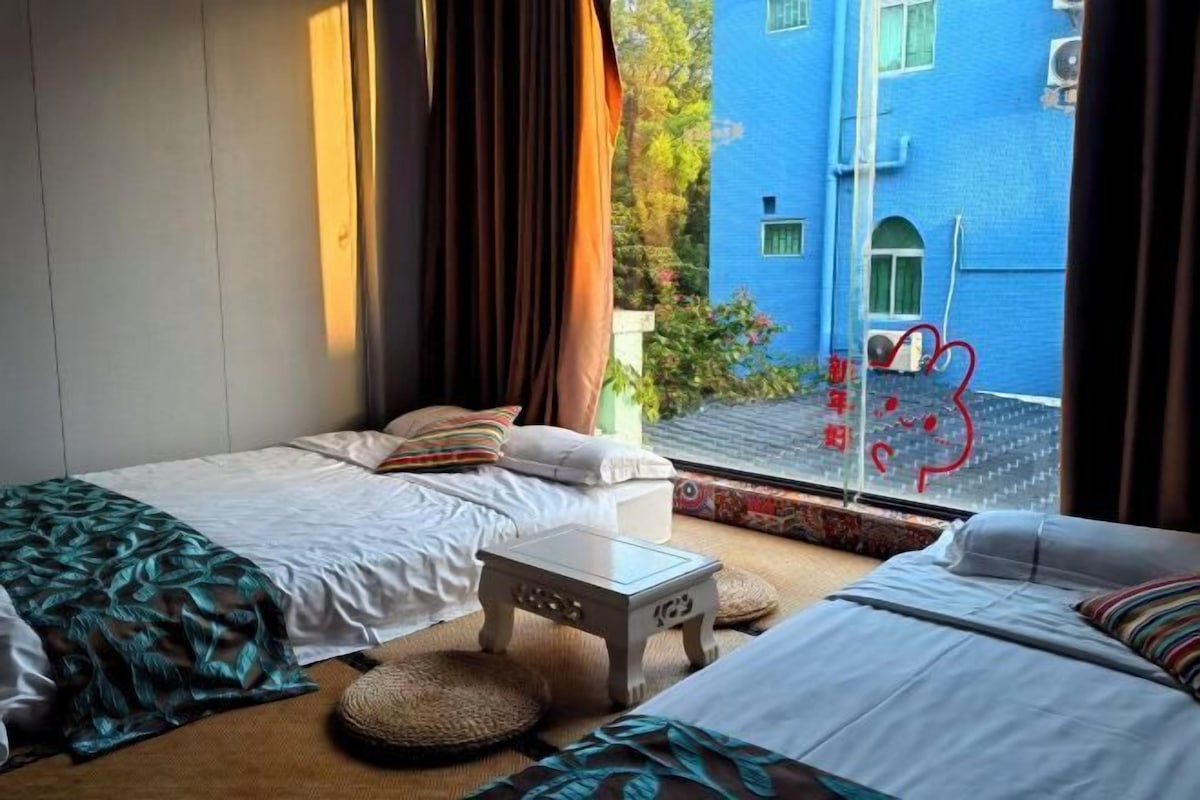 A hotel room with two beds, a small table, and a view of a blue building outside.