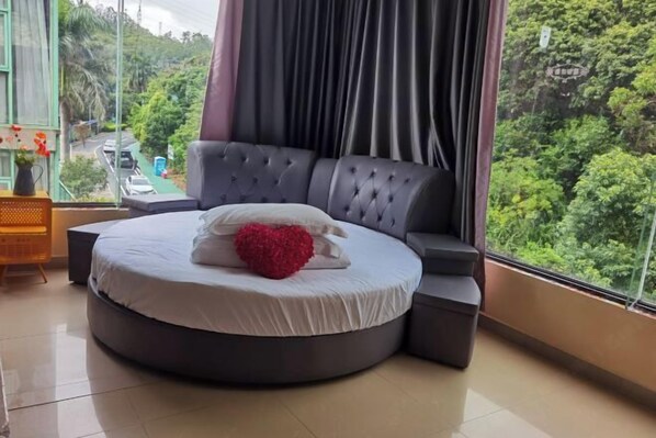 A round bed with a white mattress and a red heart-shaped pillow, a tufted headboard, and a view of a road and greenery through a window. - Yimu Huatian Inn (Shenzhen)