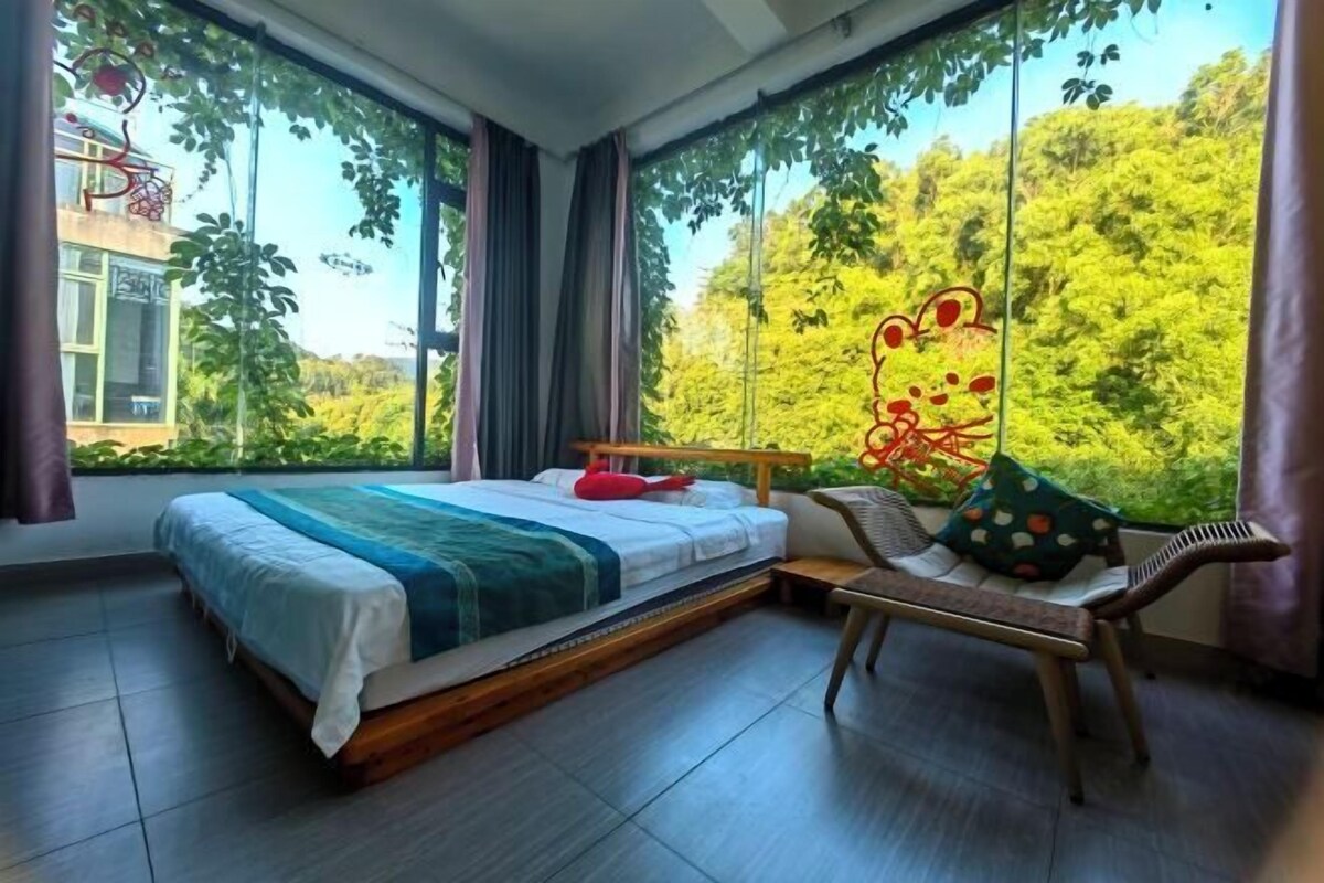 A bedroom with a large bed, a chair, and a view of greenery through large windows.