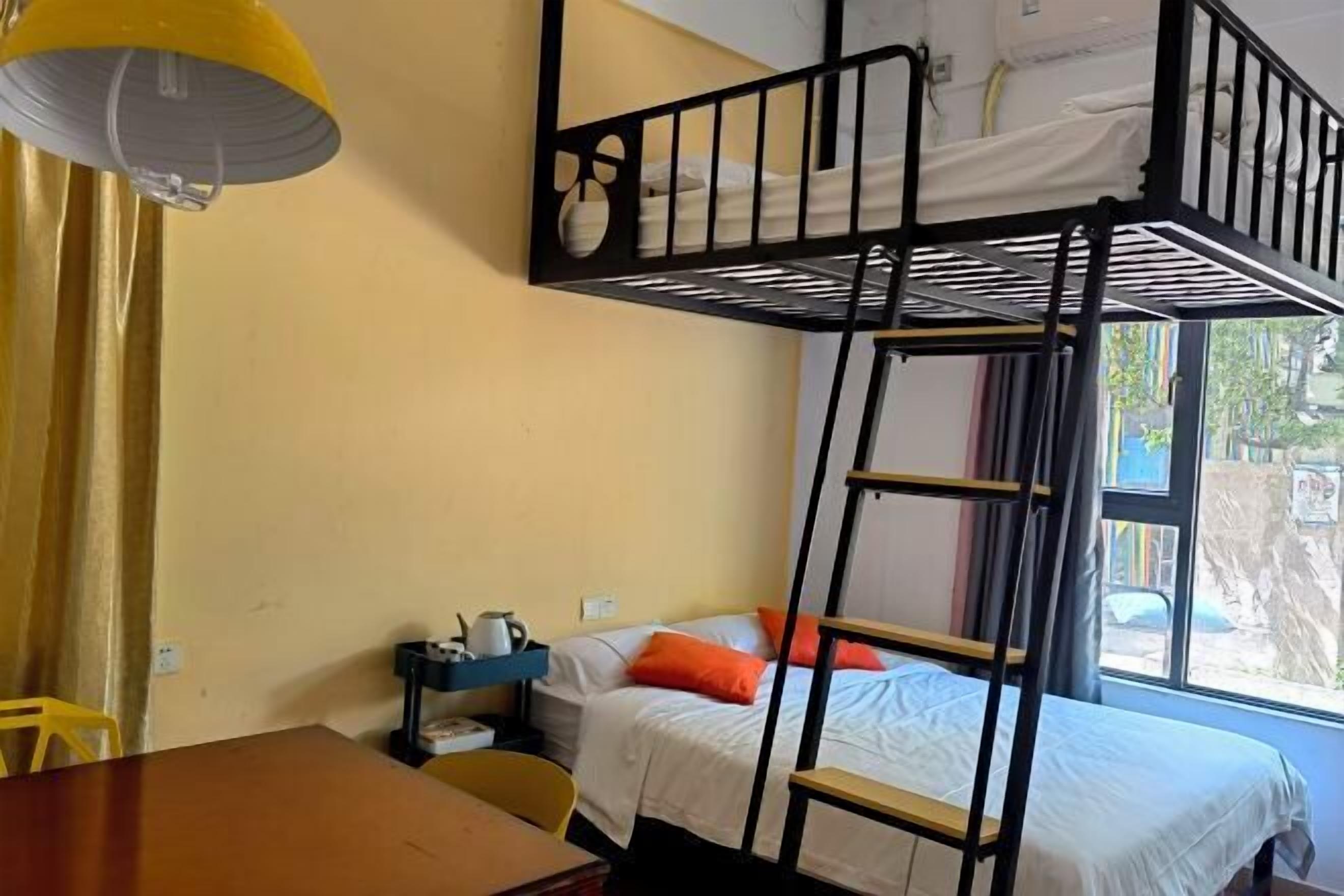 A dormitory room with bunk beds, a desk, a chair, a lamp, and a window with a view of the outside.