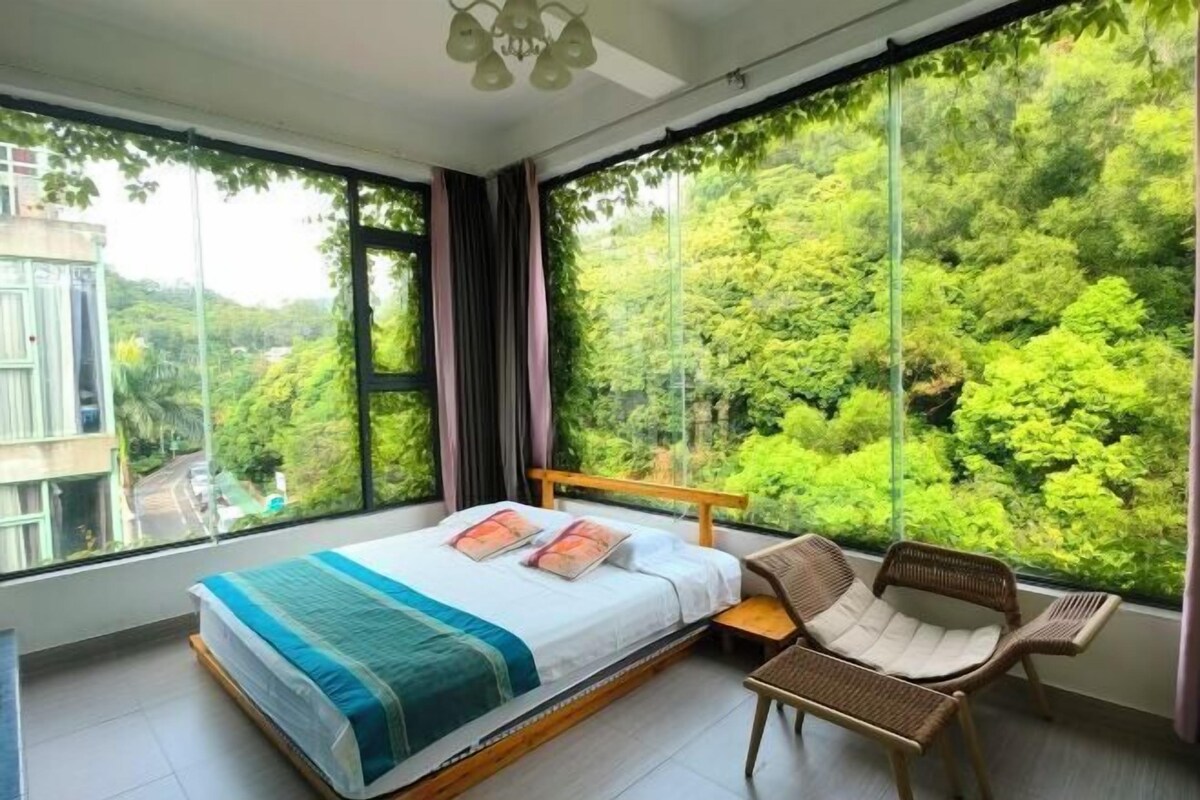 A bedroom with a large bed, a chair, and a view of lush greenery through floor-to-ceiling windows.