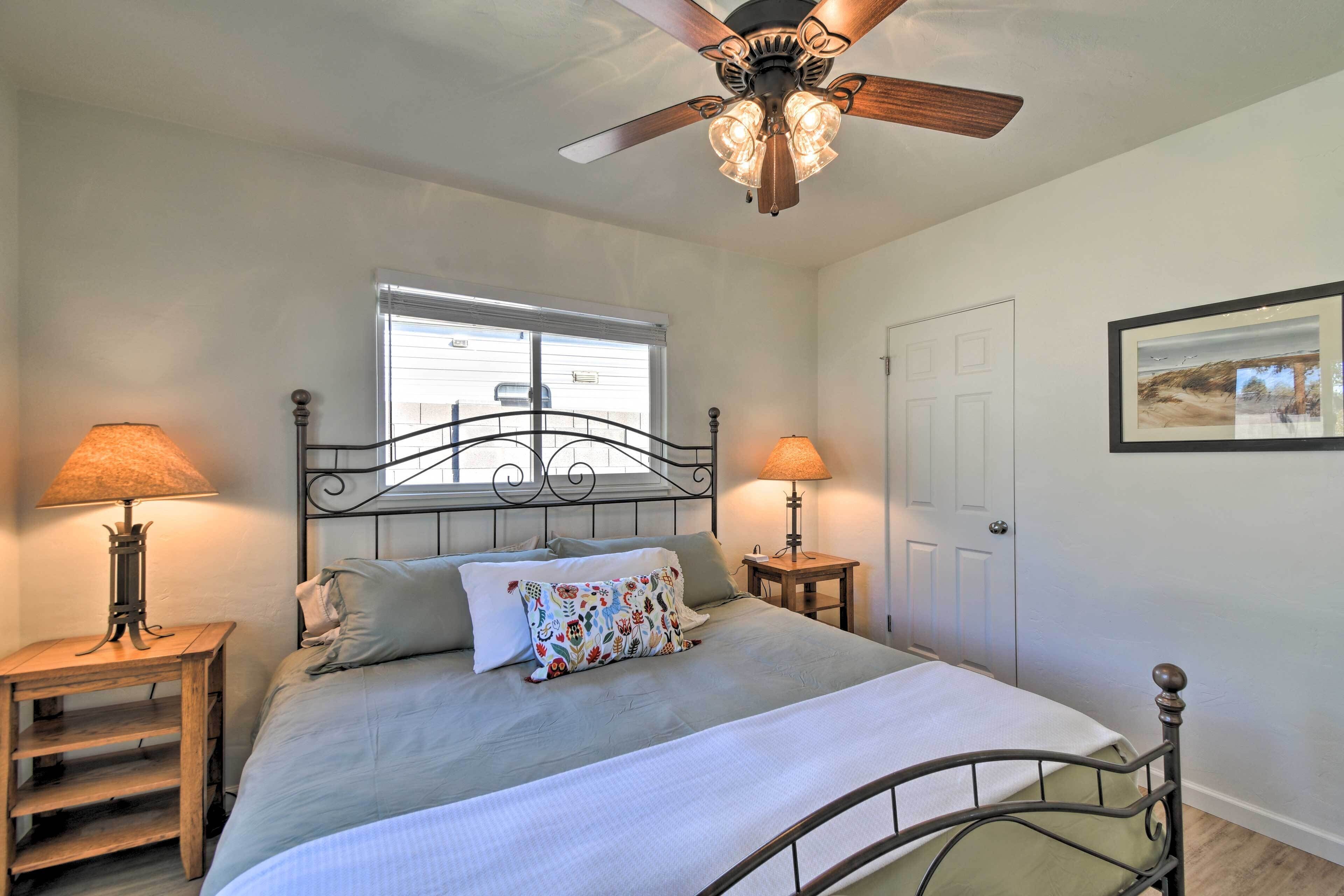 A bedroom with a bed, two bedside lamps, a ceiling fan, a window, and a door.