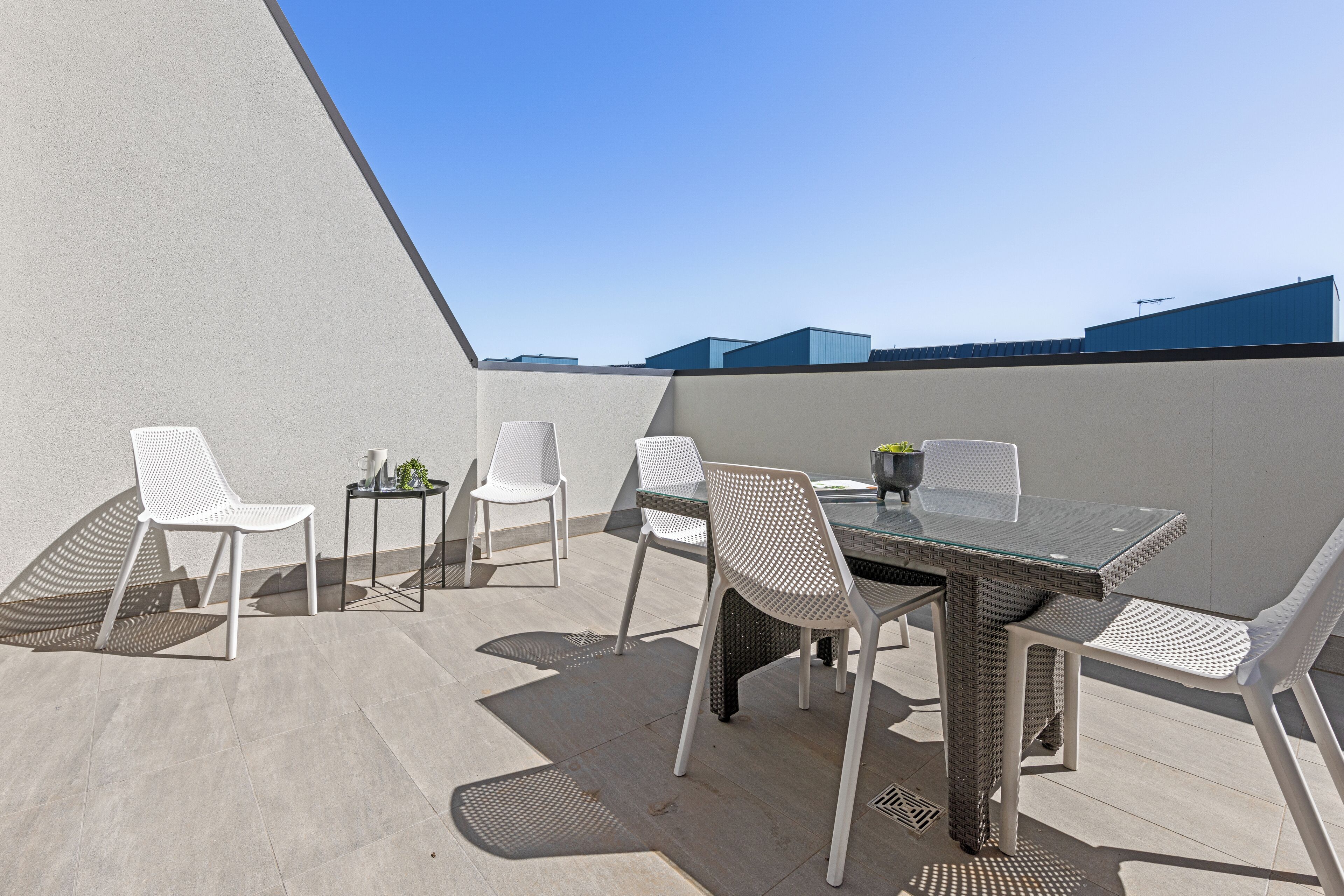 Une terrasse sur le toit, avec une table en verre et des chaises blanches, entourée d’un petit muret et surplombant un ciel dégagé.