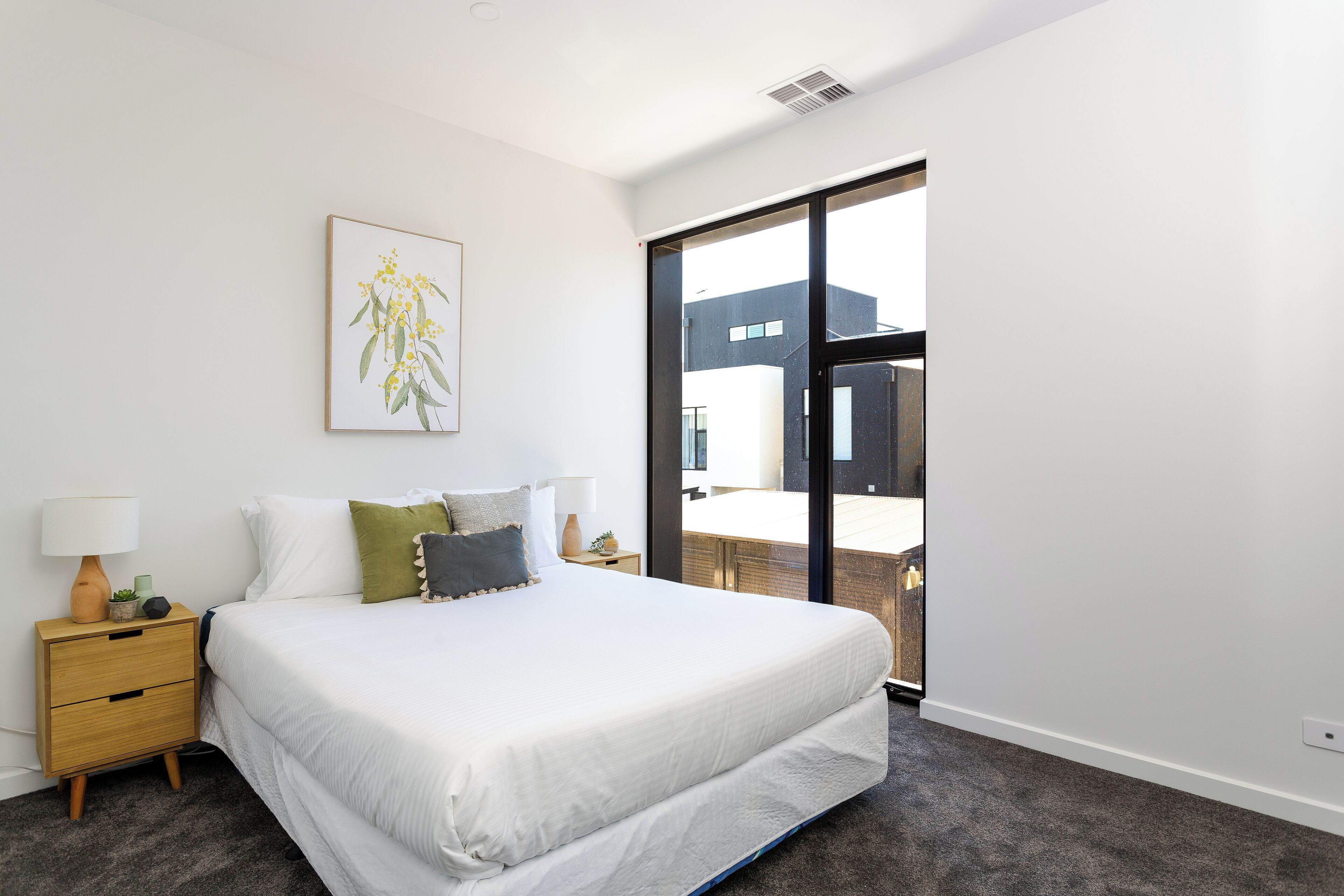 A modern bedroom with a large bed, a nightstand with a lamp, a framed picture on the wall, and a view of a building through a sliding door.