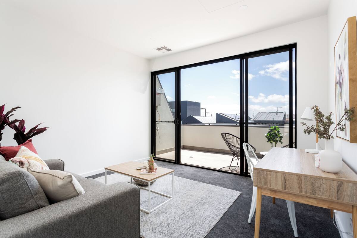 A modern living room with a grey sofa, a small wooden coffee table, and a dining area with a white table and chairs. Large windows offer a view of the cityscape.