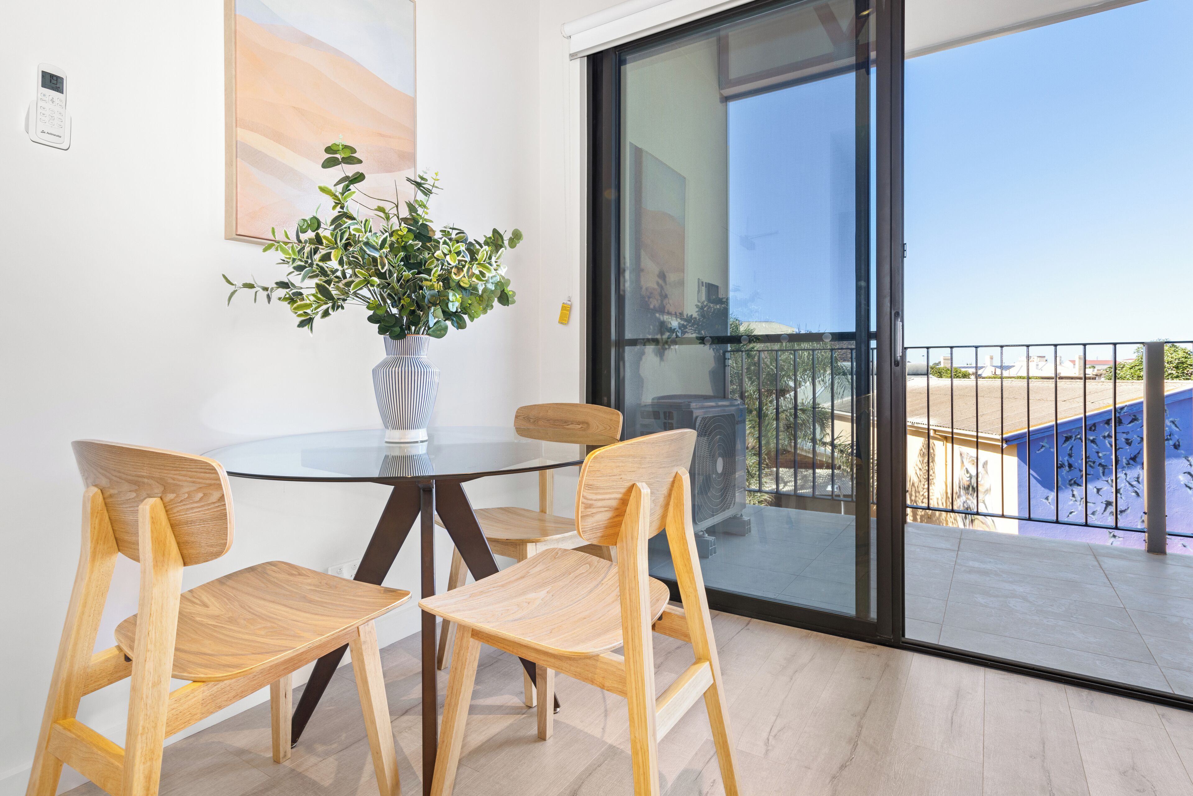 A modern room with a glass table and wooden chairs, a potted plant, and a view of the outside balcony.