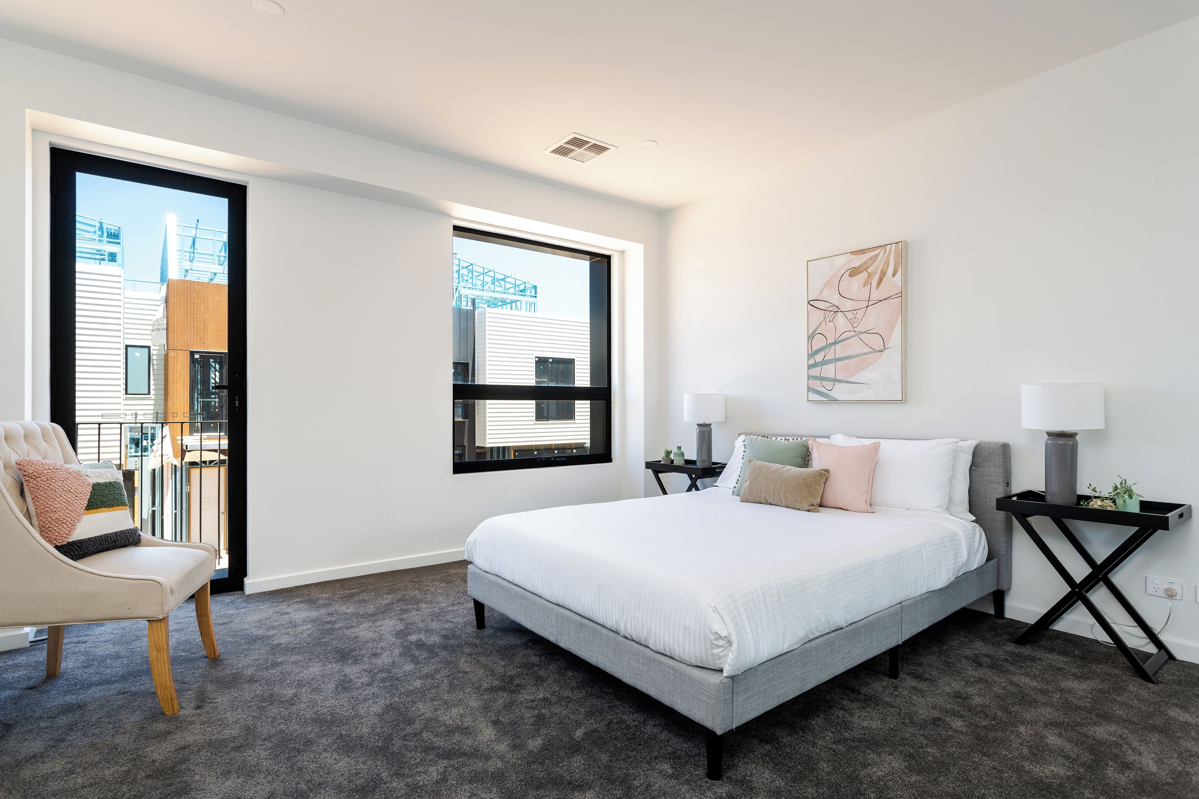 Ein modernes Schlafzimmer mit einem großen Bett, einem Sessel, einem Nachttisch und einem Fenster mit Blick auf Gebäude.