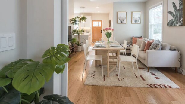 A modern living room with a dining area, featuring a wooden dining table, white chairs, a sofa, and large green plants. - Airy Downtown 1BR w/ Parking (Princeton)
