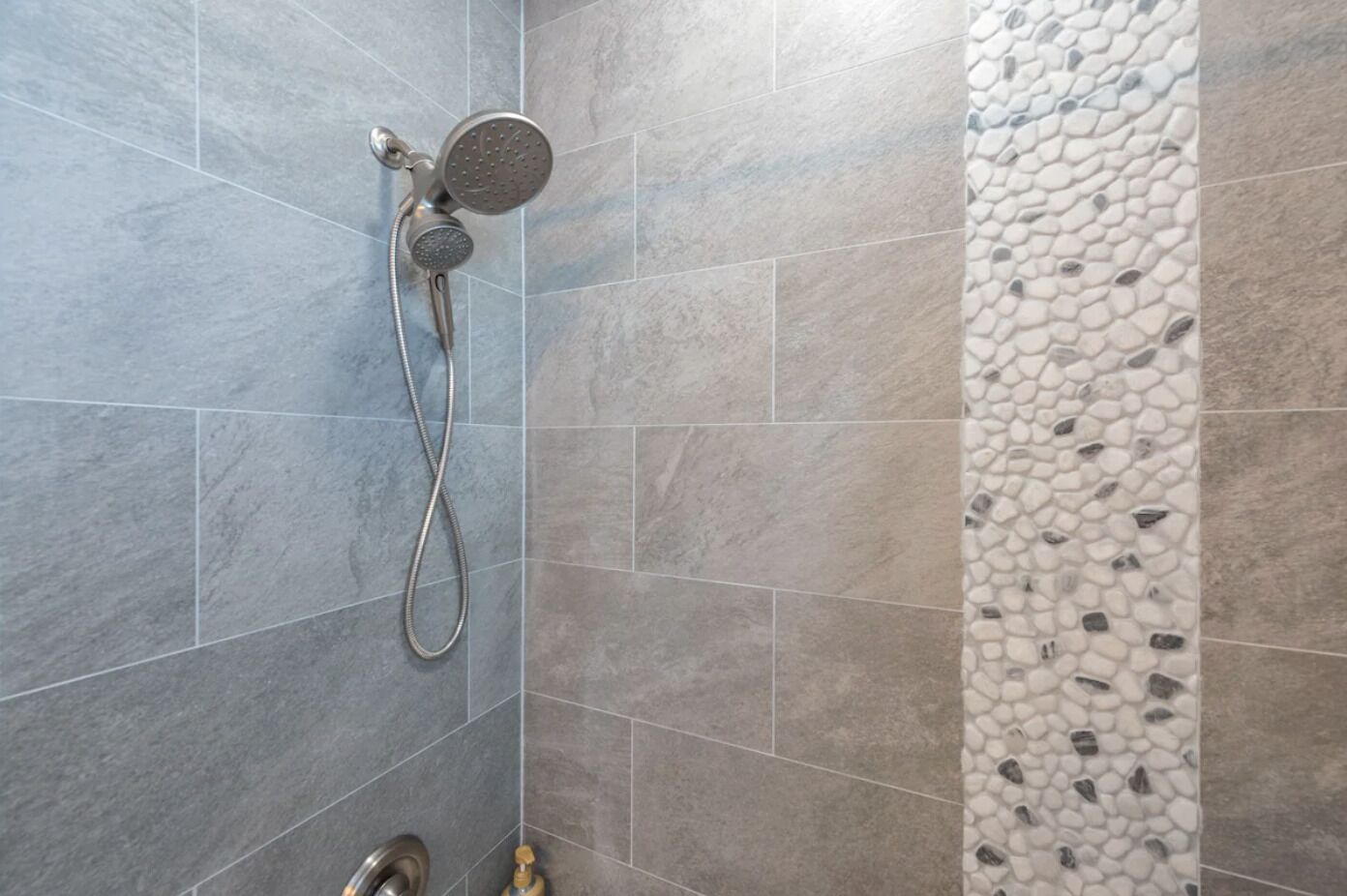 A shower with a handheld showerhead and a wall-mounted faucet, tiled with large gray tiles and a pebble-patterned accent wall.