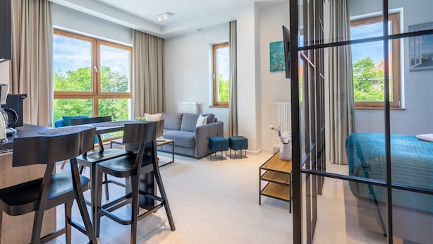 Una sala moderna con sofá, mesa de centro y comedor. Amplias ventanas con cortinas, una cama con cubierta azul y vistas a zonas verdes.