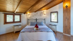 A bedroom with a wooden ceiling, two bedside tables, a bed with a white bedspread, two pillows, a nightstand with a lamp, and a window with a view of greenery. - Torrox 006 (Torrox)