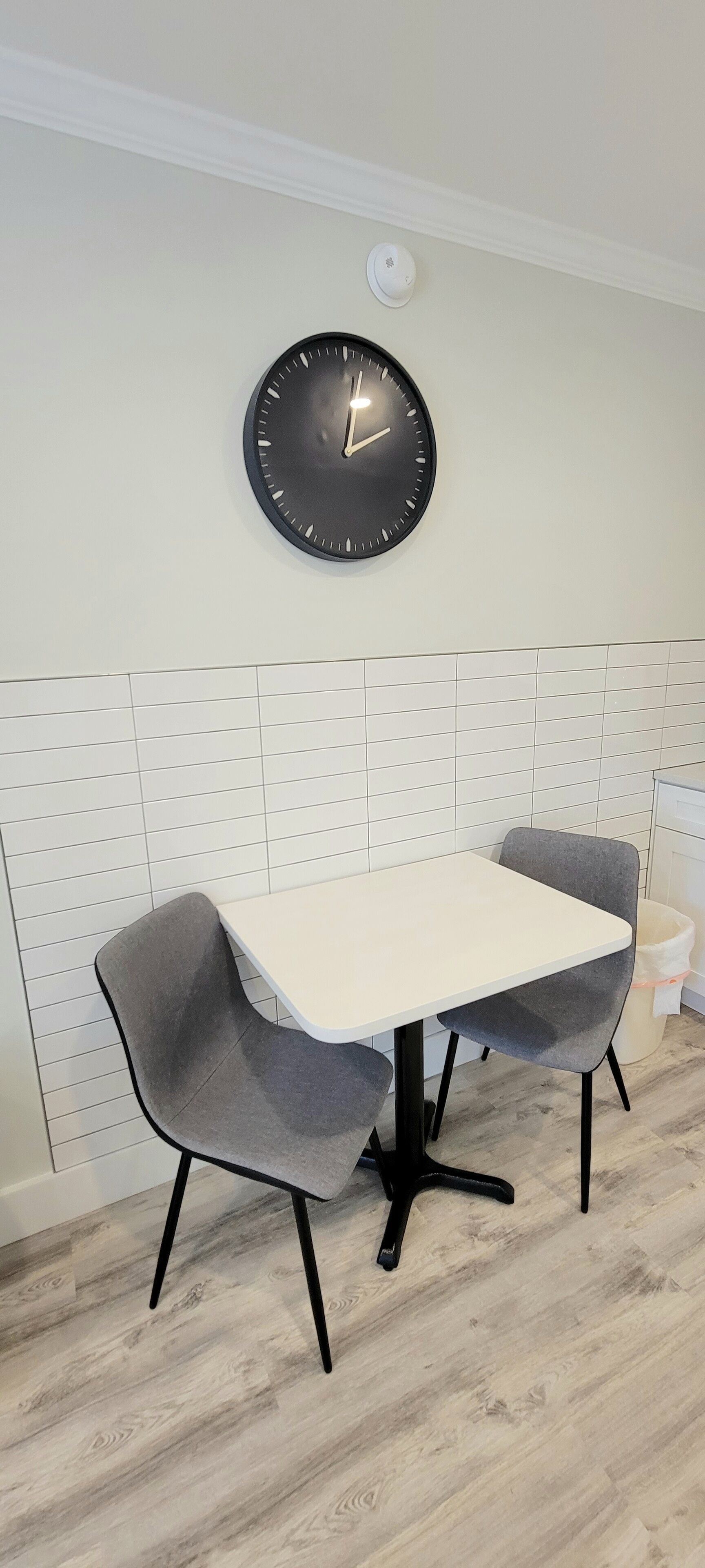 Een eetruimte met een witte tafel en twee grijze stoelen, een zwarte wandklok en een witte betegelde muur.