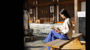 Une personne est assise sur un banc en bois, lisant un livre, dans un cadre japonais traditionnel caractérisé par une architecture en bois et des escaliers en pierre.
