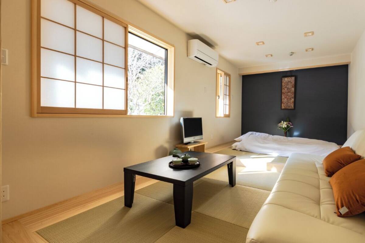 A room with tatami flooring, a bed with white bedding, a sofa with orange cushions, a television, and a small table with a decorative item.
