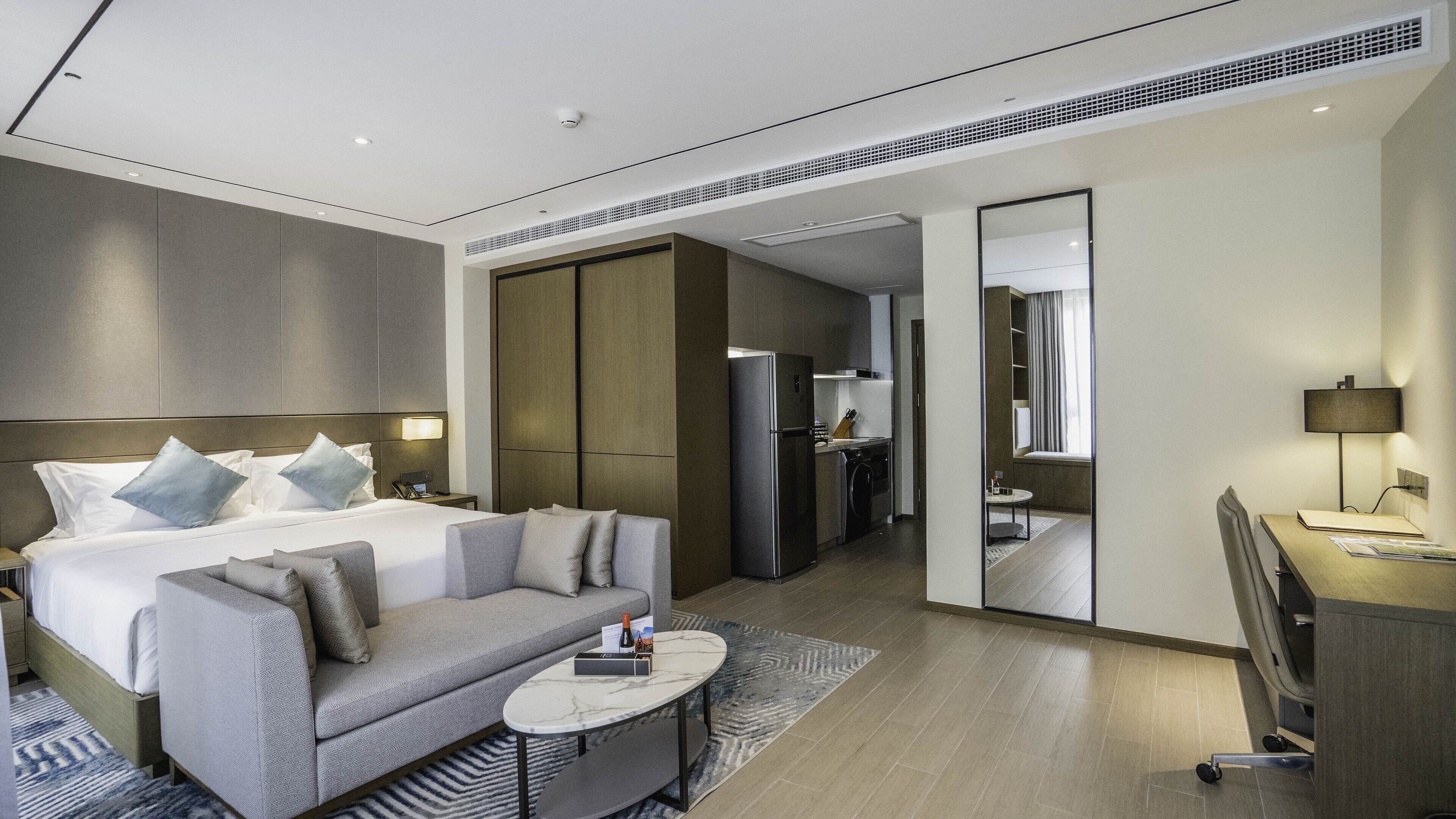 A modern hotel room with a large bed, a sofa, a small table, a desk, and a kitchen area.