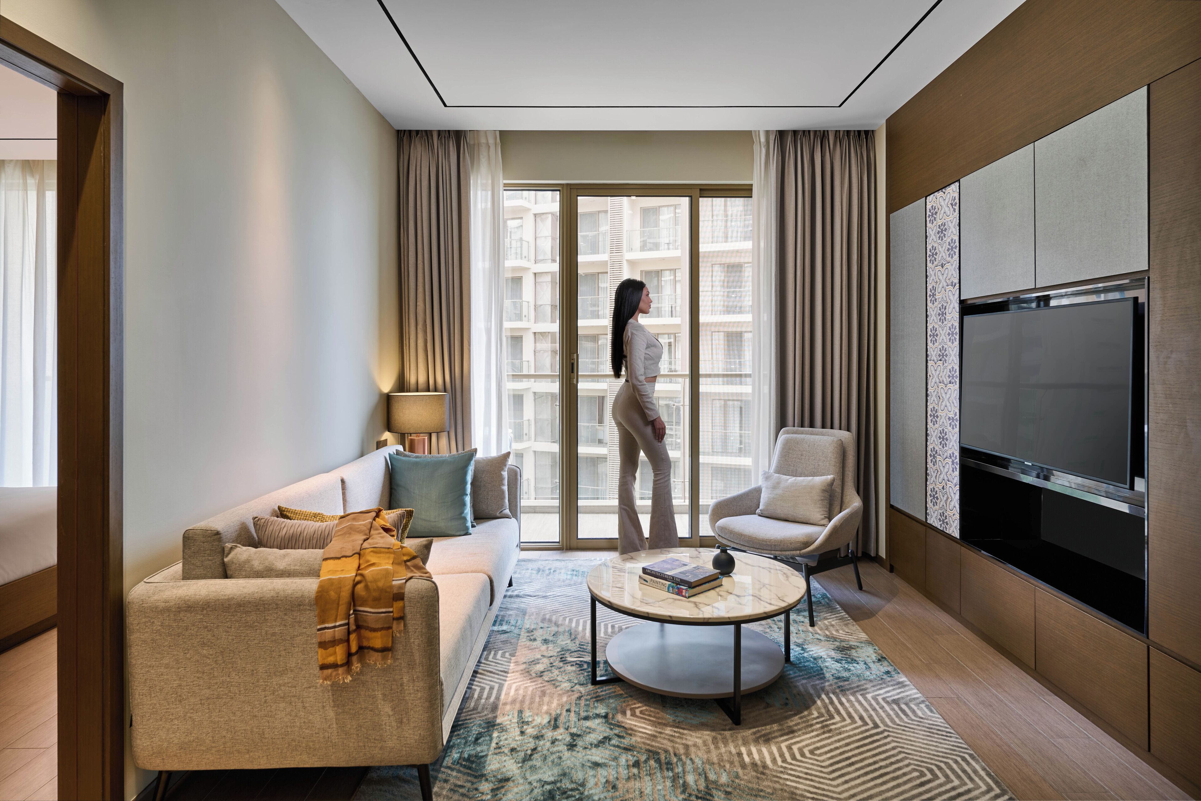A modern hotel room with a sofa, armchair, and a round coffee table. A large TV is mounted on the wall, and there is a sliding glass door leading to a balcony.